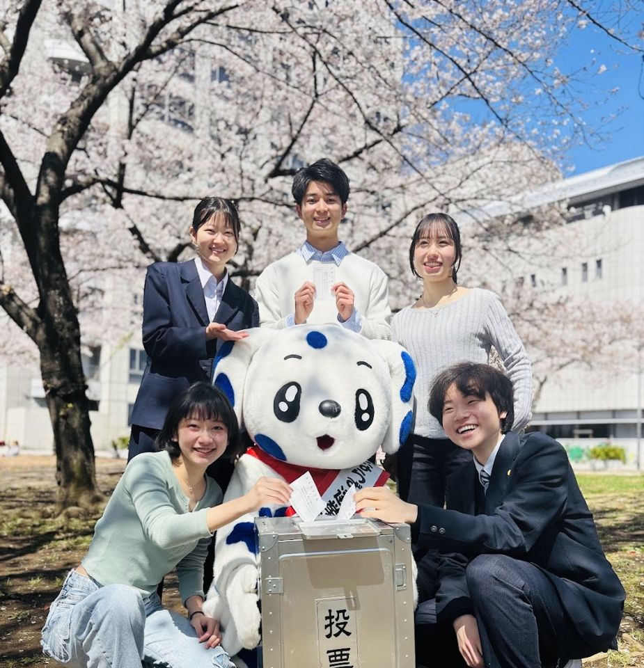桜の下、足立区のマスコットと投票箱を囲んで笑うメンバー