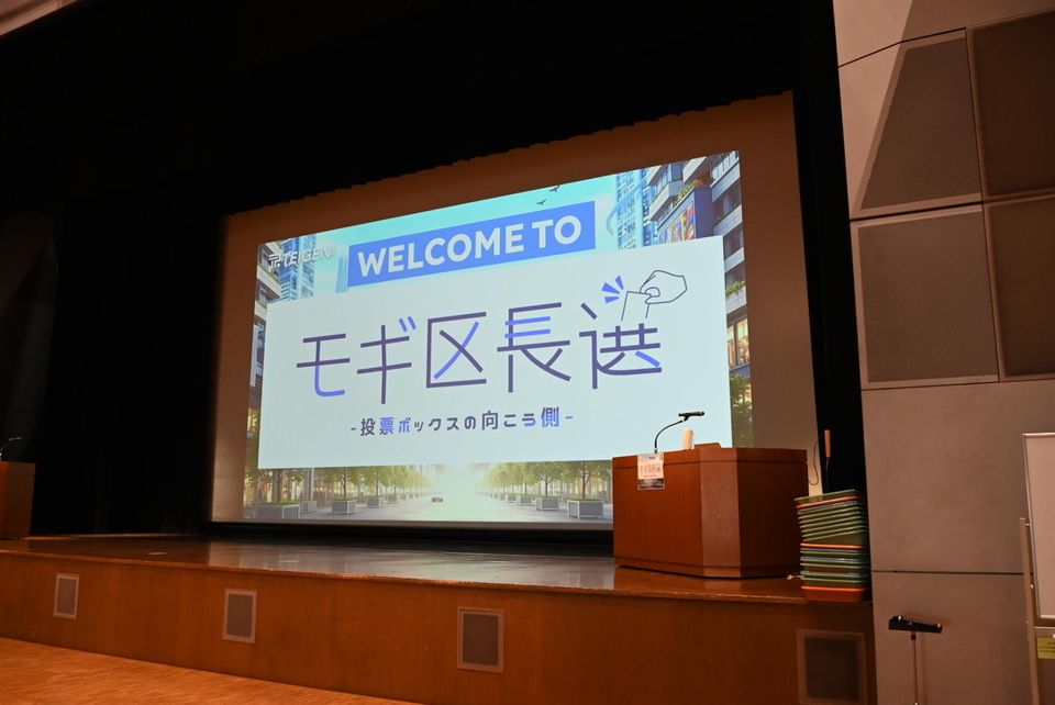 開会前のホール。スクリーンに「WELCOME TO モギ区長選」が映る