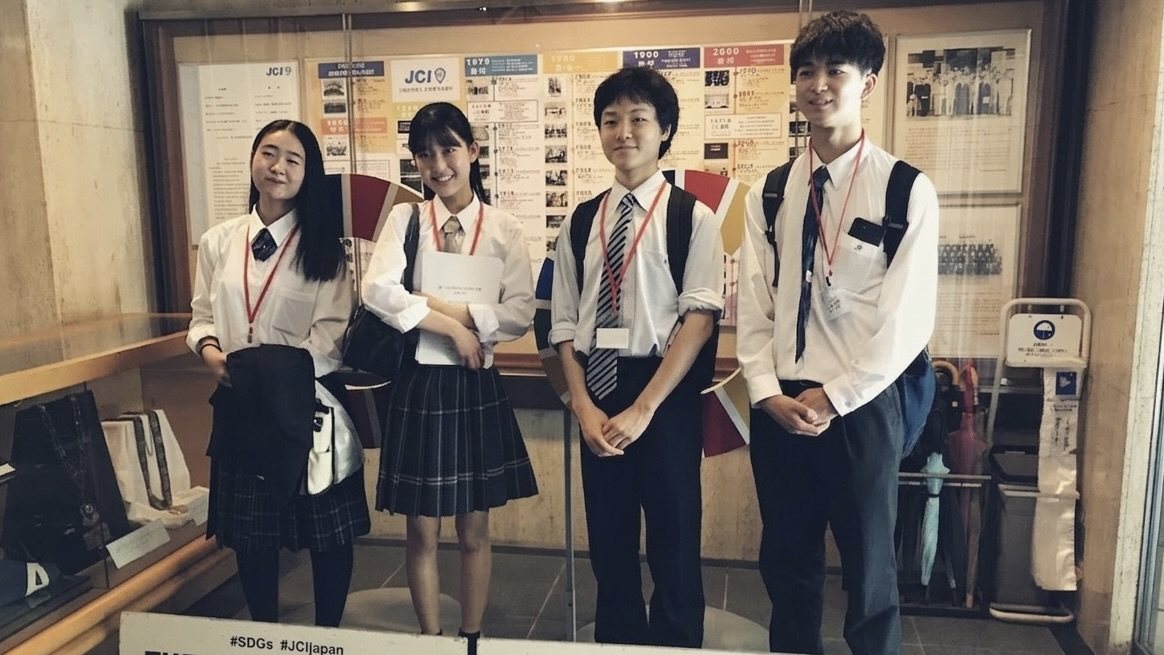 日本青年会議所の展示パネルの前に並ぶ、制服姿の創設メンバー4名