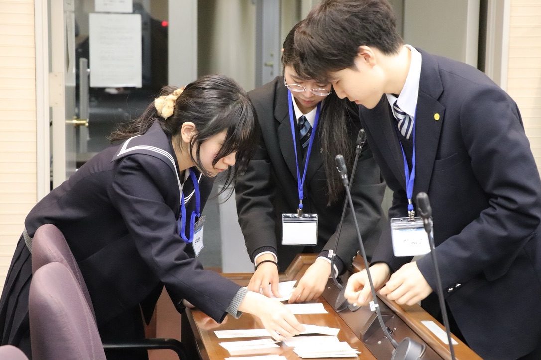 開場前の会場で、3人のメンバーが机に向かい名簿と当日資料を仕分けている様子
