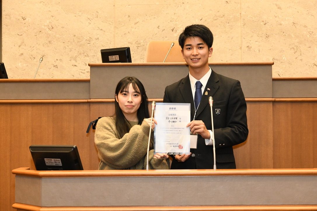 議場で、TEIGEN JP の表彰状を受賞者と並んで掲げる代表 堀井